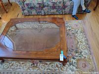 Top-down photo of wooden coffee table with beveled glass top on a patterned rug near floral couch seating.