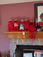 Five red plush Elmo toys arranged on a wooden mantle, varying sizes and some with clothing.