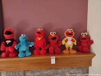 All six plush toys in a row on a wooden ledge, five red Elmo toys and one blue Cookie Monster toy.