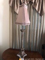 Full view of one lamp showing the entire height, detailed pleated pink fabric shade, and curved plexiglass base section on a wooden table in front of closed window blinds.