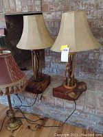 Pair of two matching table lamps with beige bell-shaped shades and natural wood twig style bases on wooden platforms, cords visible.