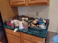 Wide view of assorted mugs, glassware, and a metal tray with kitchen utensils on green counter with wooden cabinets.