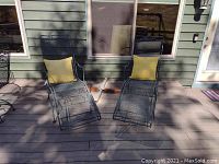 Two metal chaise lounges placed outside on a wooden deck next to a house wall and windows. Both have yellow cushions on the backrests.