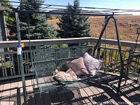 Full view of green metal mesh porch swing on deck with cushions and other items on seat, showing overall size and structure.
