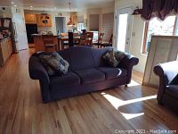 Eggplant colored sofa positioned in a living room with light wood flooring and natural sunlight. Two patterned throw pillows placed on the arms.