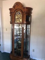 Full front view of oak tall case clock showing arched bonnet top, glass front door and brass weights