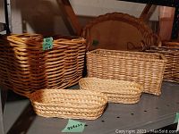 Photo showing five woven baskets and tray arranged on shelving: large square basket, basket with handle, small rectangular with lid, two small oval baskets, and large scalloped tray in background.