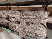 Four stacked Mirabel patio seat cushions showing leaf patterned woven olefin fabric in neutral tones, zippered sides, and thick padding.