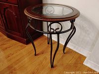 Full view of the round end table showing the glass top, wood frame, and decorative metal legs.