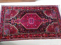 Top view of small rectangular hand woven rug showing central medallion and surrounding geometric and floral patterns in red, blue, and black.