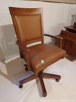 Front-side view showing the chair, leather upholstery in cognac with visible wear on seat, maple wooden armrests and frame with caster wheel base.