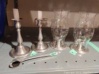 Full view of two pewter hurricane candle holders with glass chimneys, two pewter candlesticks, and one pewter candle snuffer arranged on shelf.