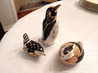 Three bird paperweights on white tablecloth: a standing penguin with cobalt blue and gold detailing, a small quail and a wren, both with intricate feather decoration in gold, blue, and orange.