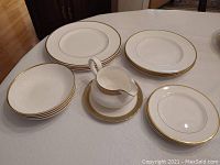 Photo of the dinner plates, bowls, side plates, and gravy boat with stand arranged on the table showing gold trim detailing and white porcelain.