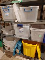 Shelf displaying multiple clear plastic storage bins of various sizes with blue handles, plus a yellow bin at the bottom right.