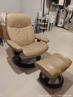 Front-left three-quarter view of recliner and ottoman showing latte leather upholstery and circular walnut base