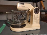 Sunbeam Mixmaster cream-colored stand mixer with 2 clear glass mixing bowls nested together on the base, metal beaters inside the bowls, rotary speed control dial visible on side.