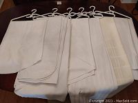 Set of six white and off-white table linens and table pads hung over plastic white hangers showing varied woven textures and patterns.