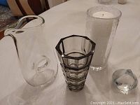 Group photo of all four items including large clear glass decanter, tall vase with candle, small faceted vase, and crystal paperweight on white tablecloth.