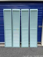 Four tall antique architectural salvage shutters painted light blue, each approximately 16 inches wide by 80 inches tall, standing side by side against a blue roll-up door.