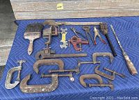 Photo of antique wooden and metal hand tools on a blue padded surface including large paintbrush, pipe wrench, several clamps, pliers, and hand drill.