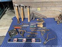 Lot of antique hand tools spread on a blue padded blanket including hammers, combination squares, hand planes, chisels, pipe wrenches, tape, level, and dividers.