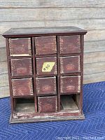 Front view of a small antique wooden cabinet with 11 small square drawers in red paint, showing wear with some drawers missing.