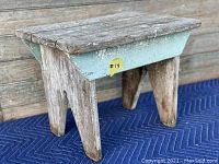 Side view of antique wooden milking stool showing three legs and faded turquoise painted apron with wear and chipping.