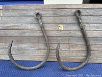 Two large antique metal hooks resting on wood background, showing full front view, rusted with loop and hook ends.