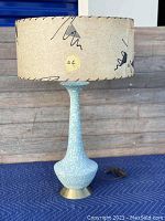 Full view of the lamp showing blue textured ceramic base with white speckles and original beige paper lampshade with abstract black scribble patterns.