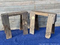 Pair of wooden antique miniature saw horses placed side by side on a blue padded surface with wooden wall background.