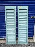 Full view of the pair of antique wooden shutters in light blue paint leaning against a blue background.