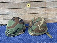 Pair of woodland camouflage covered helmets shown from side view, placed on a wooden floor with a blue padding underneath.