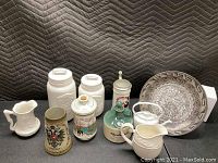 Photo of lot showing multiple ceramic bar mugs, white canisters, white pitchers, and a decorative plate on a black surface with quilted background
