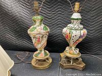 Two Capodimonte Italian porcelain table lamps side by side on black quilted fabric. Both lamps have hand-painted floral and scrollwork motifs with vintage fabric lamp shades. One lamp is missing the finial on top.