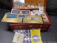 Open wooden chest with map interior showing penny folder, silver certificate, stamp holder, cloth pouch and capsule coins