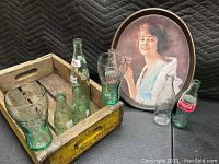Full view of wooden crate containing six Coca-Cola bottles, two Coca-Cola glasses beside it, and a Coca-Cola metal serving tray behind them. Condition is worn and vintage.