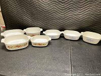 Seven white Corningware ceramic baking dishes arranged in two rows showing two different floral patterns. Four dishes with a multicolor French pattern and three dishes with a blue flower pattern.