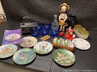 Photo of the entire lot showing decorative plates, blue glassware, cast-iron mini stove, doll figurine, and small decorative items