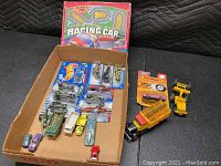 Photo showing a cardboard box with 13 Hot Wheels cars, some in original blister packages, and an assortment of loose cars and a Racing Car game box in the background.