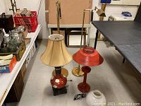 Photo showing four lamps grouped on floor: two red-orange ceramic table lamps with floral patterns, one with beige shade, the other missing shade; one floor lamp with brass/gold stand and small brass shade; and one bare lamp stand.