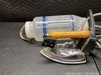 Side-angle view of the Naomoto industrial steam iron showing the metal soleplate, wooden handle, steam valve and attached water reservoir bottle with Naomoto Industrial Steam Co. label.