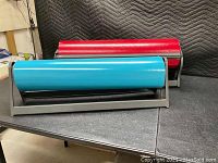 Two paper rollers with metal frames on table, one red and one blue roller tube.
