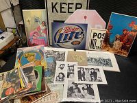 Photo showing a collection of vintage lithographs, Beatles press photos, vintage advertisements including a Miller Lite sign, and other ephemera all spread on a table.