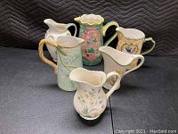 Photo of six large decorative ceramic pitchers with various floral and decorative designs shown from front view