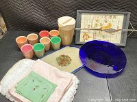 Seven colorful small cups, wooden pitcher with lid, an embroidered linen piece, framed tray with culinary illustrations, and two platters (one cream ceramic with floral design and one deep blue glass) arranged on black fabric.
