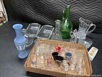 Overview photo showing all glass items on a table including shot glasses in a cardboard box, various vases, green bottle, pitcher, and trays.