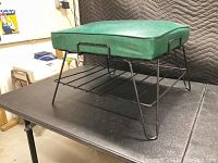 Side angle of green vinyl cushioned foot rest showing black iron wire hairpin legs and magazine rack shelf.