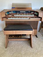 Front view of the vintage electric organ with two keyboards, control switches, wooden speaker cover, and matching bench in front.
