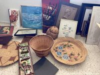 Wide angle photo showing a variety of framed artwork leaning against the wall along with woven baskets and small decorative items on the floor.